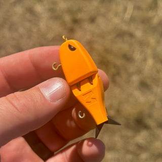 A close-up of a hand holding the Pumpkin Raptor fishing lure outdoors against a blurred tan grassy background.