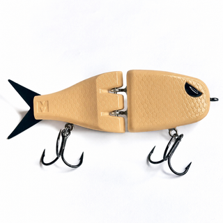 A tan fish-shaped fishing lure with a black tail, black eye detail, and two black treble hooks, photographed on a clean white background.
